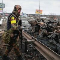 An armed man stands by the remains of a Russian military vehicle in Bucha, close to the capital Kyiv, Ukraine, Tuesday, March 1, 2022. (AP Photo/Serhii Nuzhnenko)